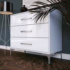 White dresser with silver legs and drawers against a dark wall with a plant and lamp.