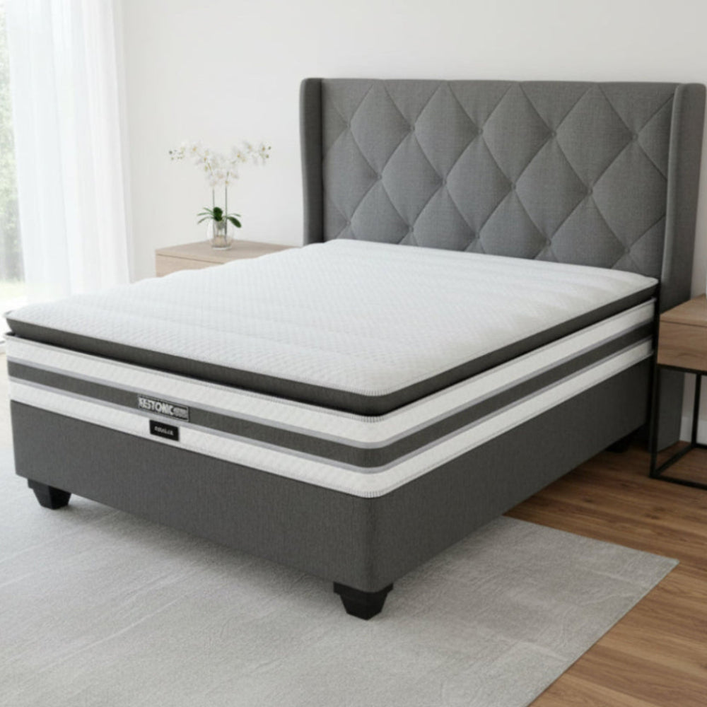 Gray and white mattress with quilted headboard in a room with a window and side table.